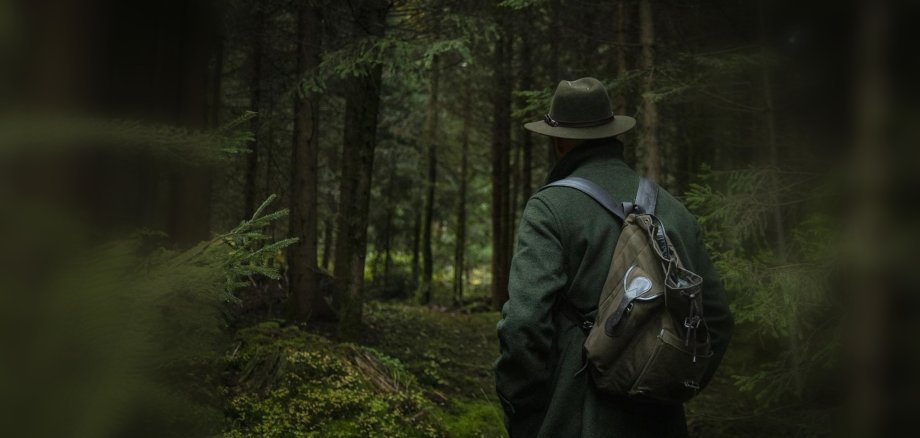 Ein Jäger schaut in den Wald hinein, man sieht ihn von hinten mit Hut und Rucksack, umrandet von Bäumen und Ästen.